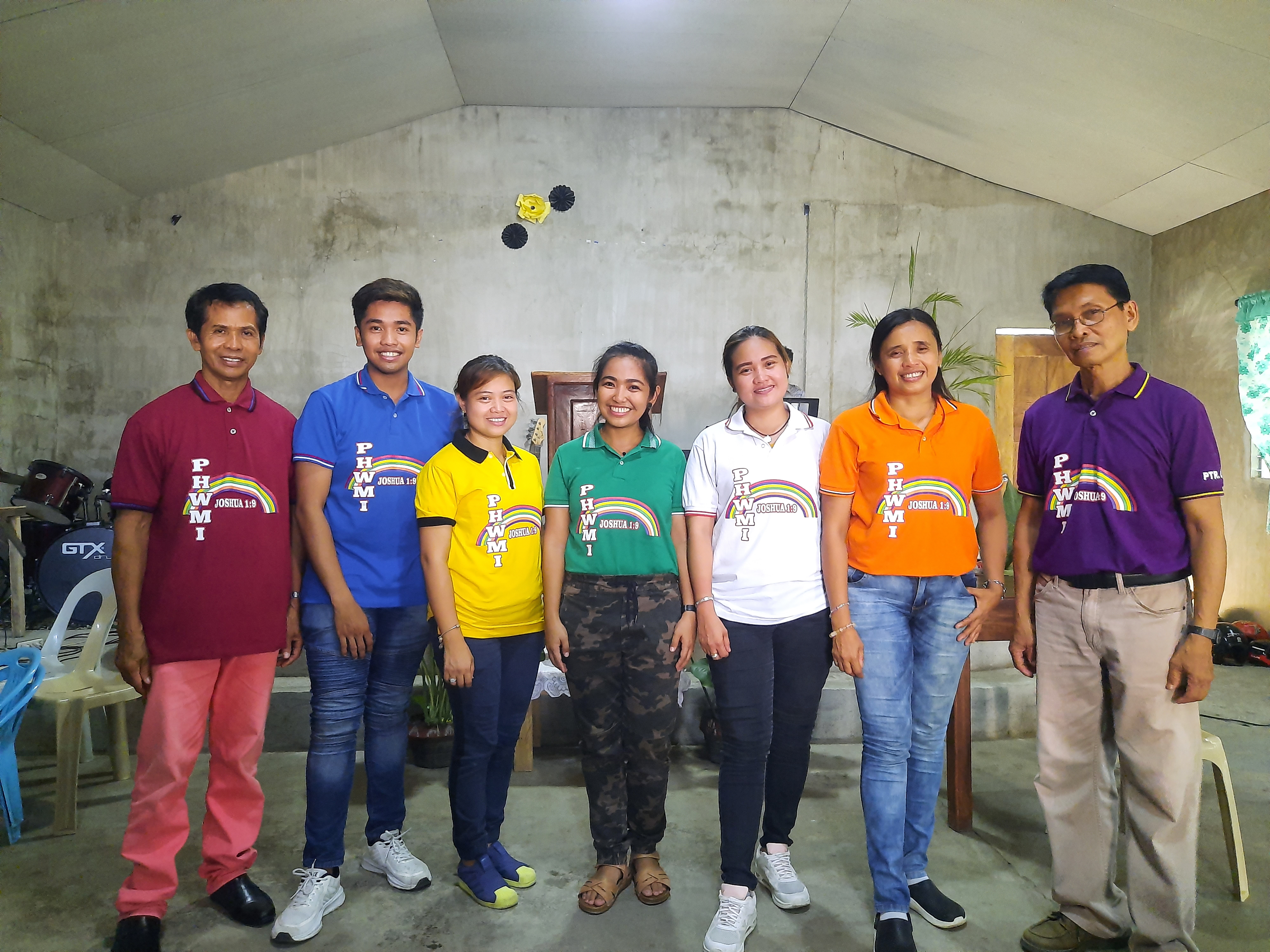 Seven PHWMI leaders standing together, each wearing a different colored shirt with the PHWMI rainbow emblem